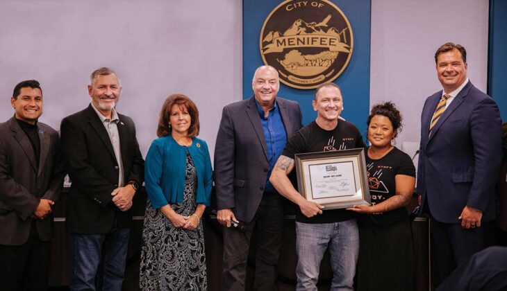 A group of seven people stands together, with two individuals in the center holding a framed certificate and smiling. The backdrop features a "City of Menifee" emblem, celebrating their achievement in Menifee's Business Spotlight.