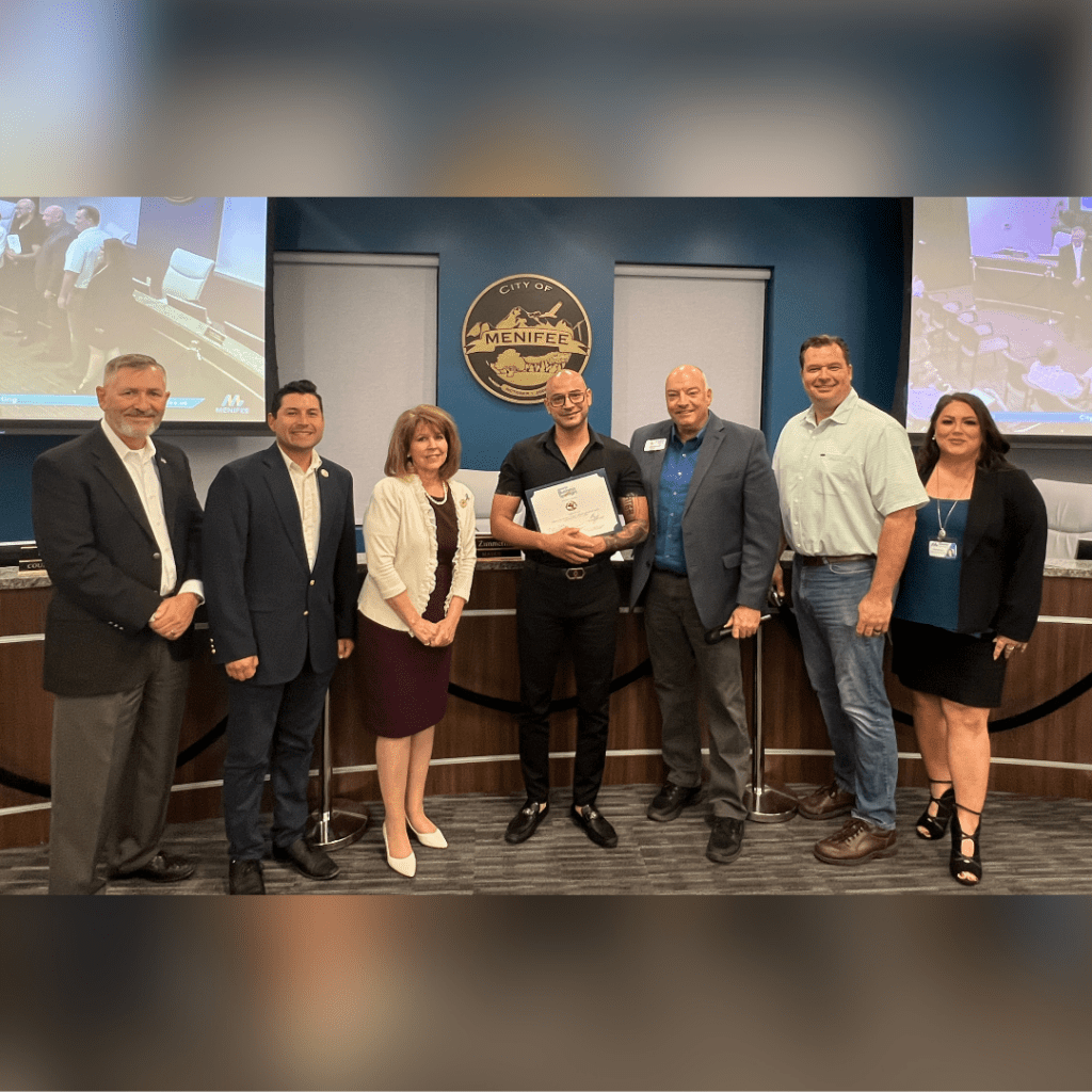 A group of seven people standing and smiling in front of a City of Menifee council chamber. One person in the center is holding a certificate celebrating the October Business Spotlight for Tacos & Tequila Grill, highlighting their success as a Menifee business.