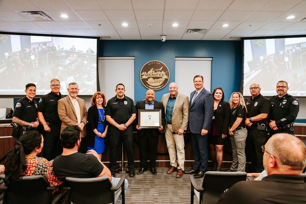 A group of people stands in a conference room, with one person holding a framed certificate. Two screens display a mirrored image of the group. The room has the City of Menifee emblem on the wall, celebrating their recognition in the Menifee Business Spotlight for May Business.