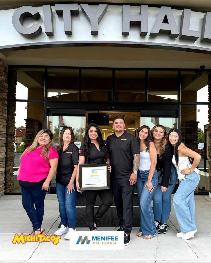 A group of people stands outside City Hall, posing with a framed certificate. The sign above reads "CITY HALL." Two logos at the bottom read "Michi Tacos" and "Menifee California," highlighting their featured Business Spotlight.