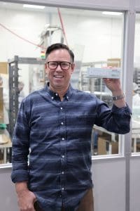 A person in a blue shirt holds a box labeled Davids Natural Toothpaste while standing in a manufacturing facility.